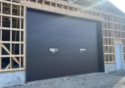 A large brown garage door with two small windows is installed upside down in a wooden building frame, with the windows positioned low to the ground. A white door is visible to the right.