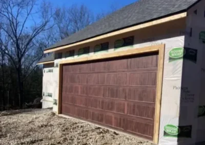 A house under construction with a large wooden garage door but no driveway, surrounded by gravel and unfinished ground, with trees in the background under a clear blue sky.