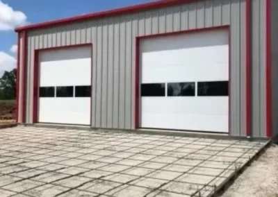 A metal building with two large garage doors stands behind a grid of steel rebar, prepared for pouring concrete, under a clear blue sky.