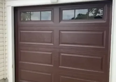 A closed brown garage door with rectangular panels and four small windows at the top, set in a white frame attached to a light-colored building exterior.