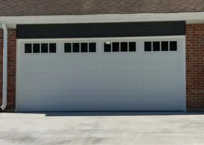 A closed white double garage door with rectangular windows at the top, set in a red brick wall with a shingled roof and a concrete driveway in front.