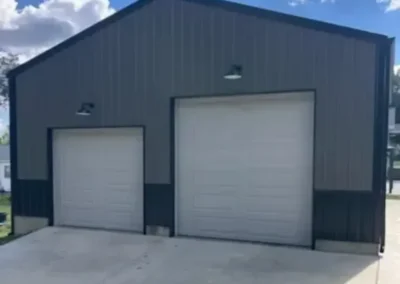 A metal building with two large white garage doors and a sloped roof sits on a concrete driveway under a partly cloudy sky.