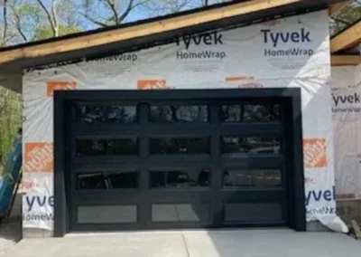 A modern glass-panel garage door installed on a house under construction, with Tyvek HomeWrap covering the exterior walls and an unfinished roof visible above. Trees and a clear sky are in the background.