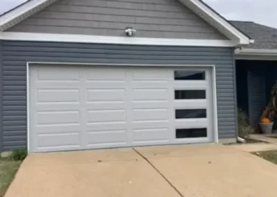 A gray house with a white garage door featuring three rectangular windows on the right side, set against a cloudy sky with a concrete driveway in front.