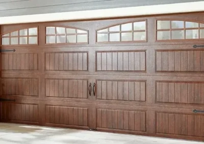 A modern house with a double wooden garage door featuring rectangular panels, decorative handles, and upper windows. Two potted plants sit on either side of the garage entrance.