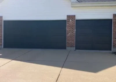 A house with three garage doors: two large black doors on the left and a smaller black door on the right, all framed with red brick and white siding above, facing a wide concrete driveway.