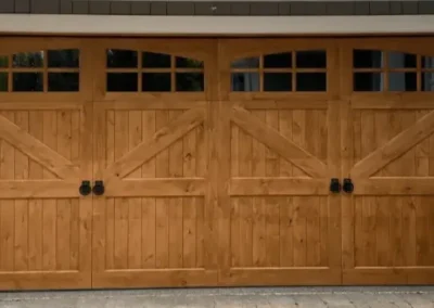 A double wooden garage door with four black handles, decorative windows at the top, and two large flower pots with orange and white flowers on either side.