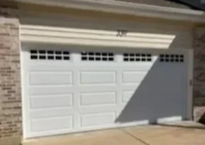 A white, double-car garage door with rectangular windows at the top is attached to a brick house. Sunlight casts a large diagonal shadow across part of the garage and driveway.