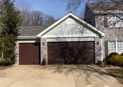A house with a three-car garage, two large brown doors and one smaller door, brick and siding exterior, a wide driveway, and tree shadows cast across the front.