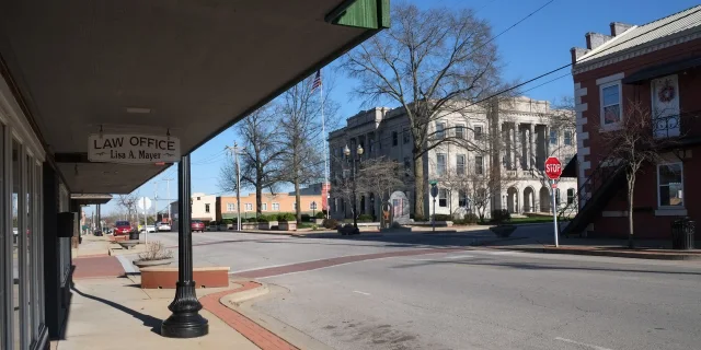 A quiet downtown street with bare trees, a law office sign, and a large historic courthouse building in the background on a clear, sunny day.