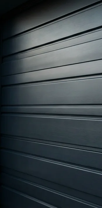 A close-up view of a modern, dark gray metal garage door with horizontal panels and a smooth, sleek finish. The lighting highlights the texture and reflective surface of the door.