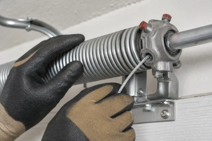 Close-up of gloved hands adjusting or repairing a metal garage door torsion spring mechanism mounted on a bracket. Close-up of gloved hands adjusting or repairing a metal garage door torsion spring mechanism mounted on a bracket.