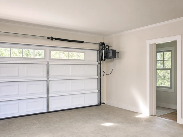 A clean, empty garage with a white automatic overhead door, beige walls, and a doorway leading to a bright room with windows. Natural light enters through the garage windows. A clean, empty garage with a white automatic overhead door, beige walls, and a doorway leading to a bright room with windows. Natural light enters through the garage windows.