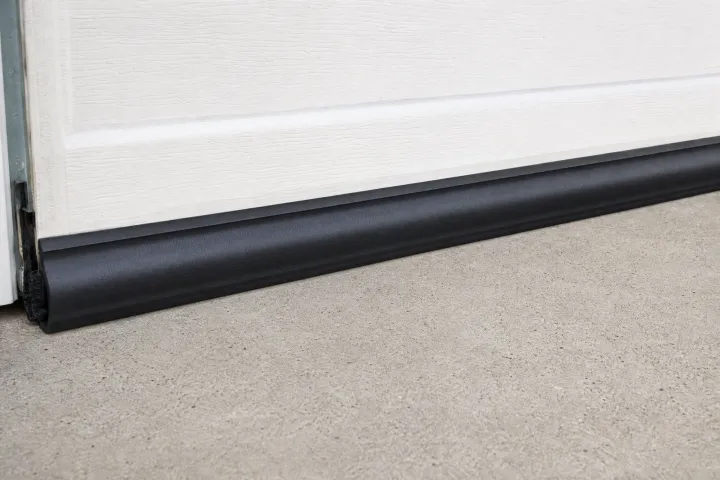 A close-up of a white garage door with a black rubber seal along the bottom edge, resting on a smooth concrete driveway. A close-up of a white garage door with a black rubber seal along the bottom edge, resting on a smooth concrete driveway.