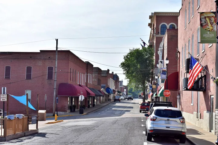 A small-town street with brick buildings, parked cars, storefronts with awnings, a stop sign, American flags, and banners hanging from lampposts on a sunny day.