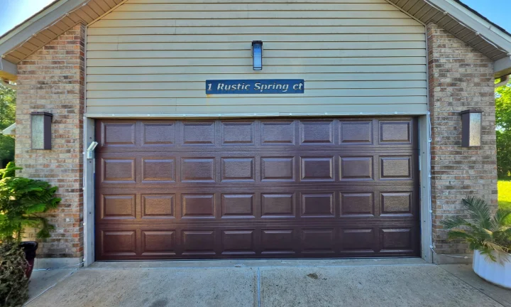A closed brown garage door on a brick house with the address 1 Rustic Spring Ct above it. Plants are on either side of the driveway.