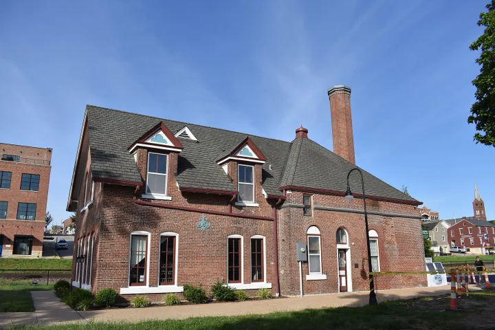 A two-story red brick building with tall windows, dormer windows, and a large chimney, set against a clear blue sky. There are other buildings and trees in the background.