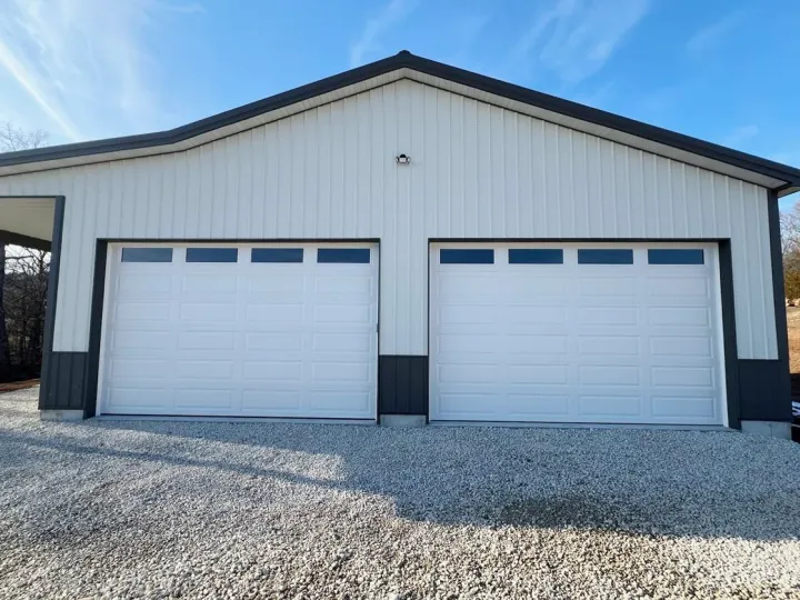 A large metal building with two white rural garage doors and small rectangular windows at the top, set on a gravel driveway under a blue sky.