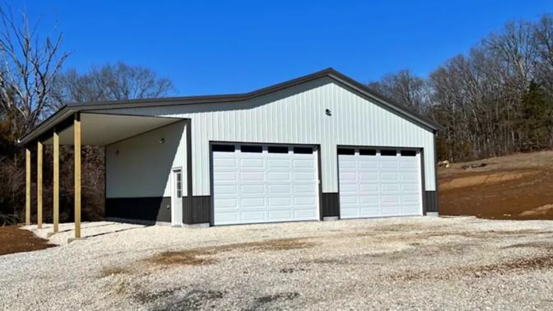 A large metal building with rural garage doors—two white garage doors and a side carport—stands on a gravel lot, surrounded by bare trees under a clear blue sky.