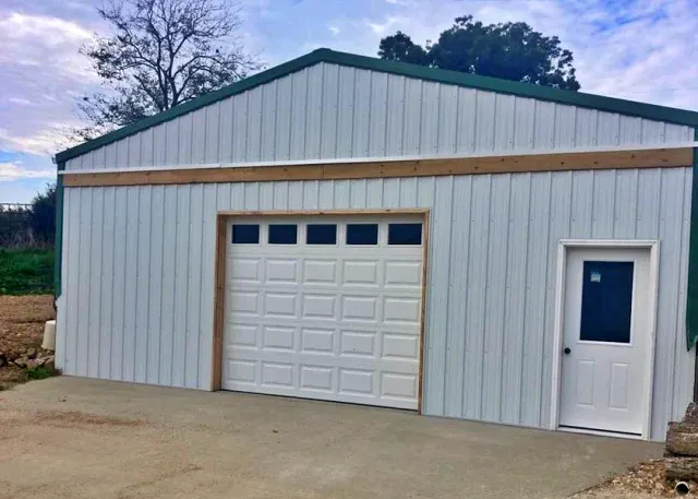 A white metal garage with a single rural garage door and a separate entry door on the right, set on a concrete driveway, with trees visible in the background.